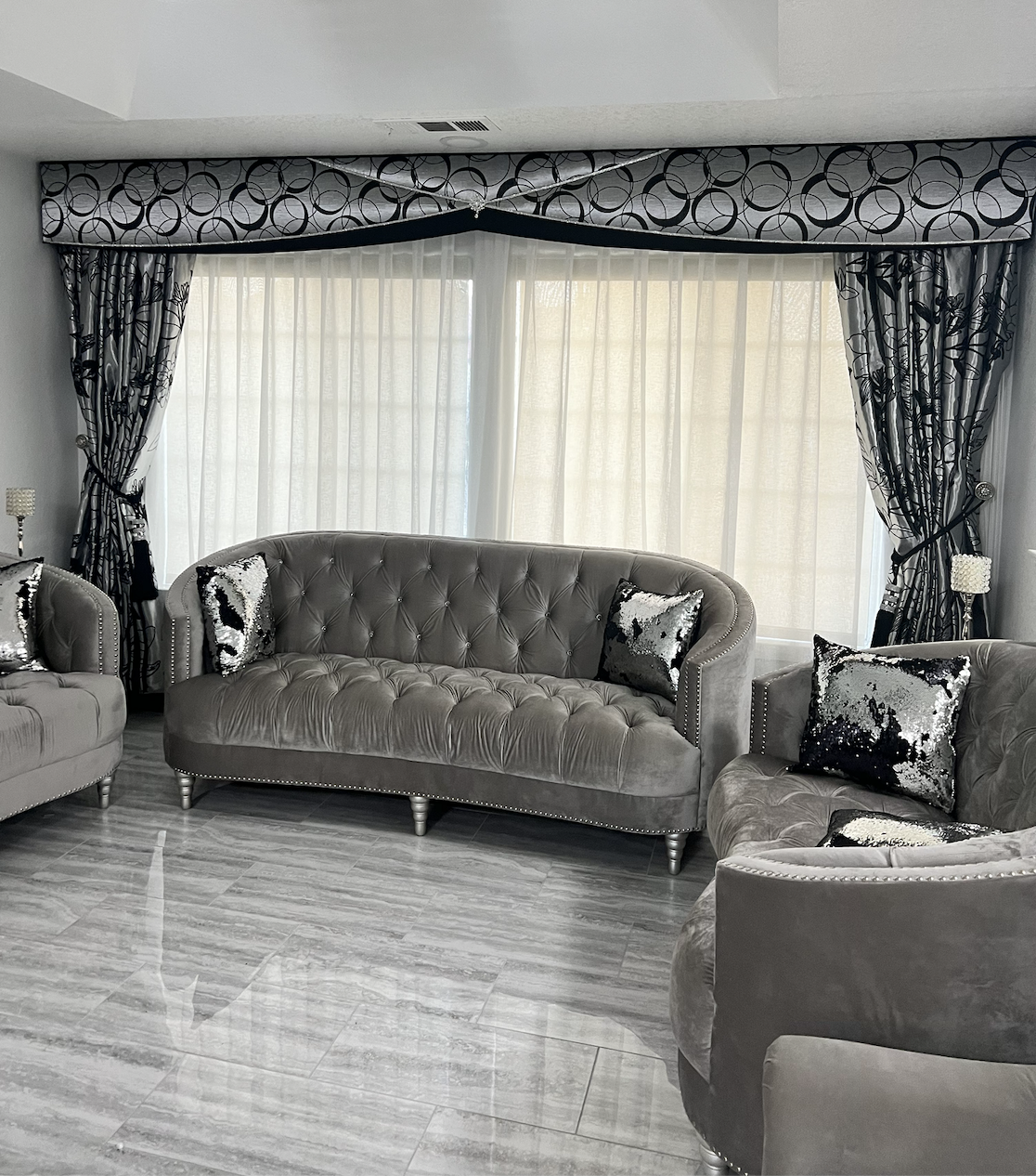 Bakersfield formal living room with a scalloped upholstered cornice box in silver and black circle-pattern fabric trimmed in crystal beadwork, spanning twin tall windows with sheer roller shades and side drapery panels in coordinating black-and-silver botanical fabric tied back with jeweled holdbacks, over a tufted grey velvet sofa and matching chairs with sequin accent pillows
