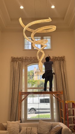 Drapery technician on rolling scaffold installing decorative top treatments at the top of a two-story arched window in a Burbank great room, with a sculptural crystal chandelier suspended overhead