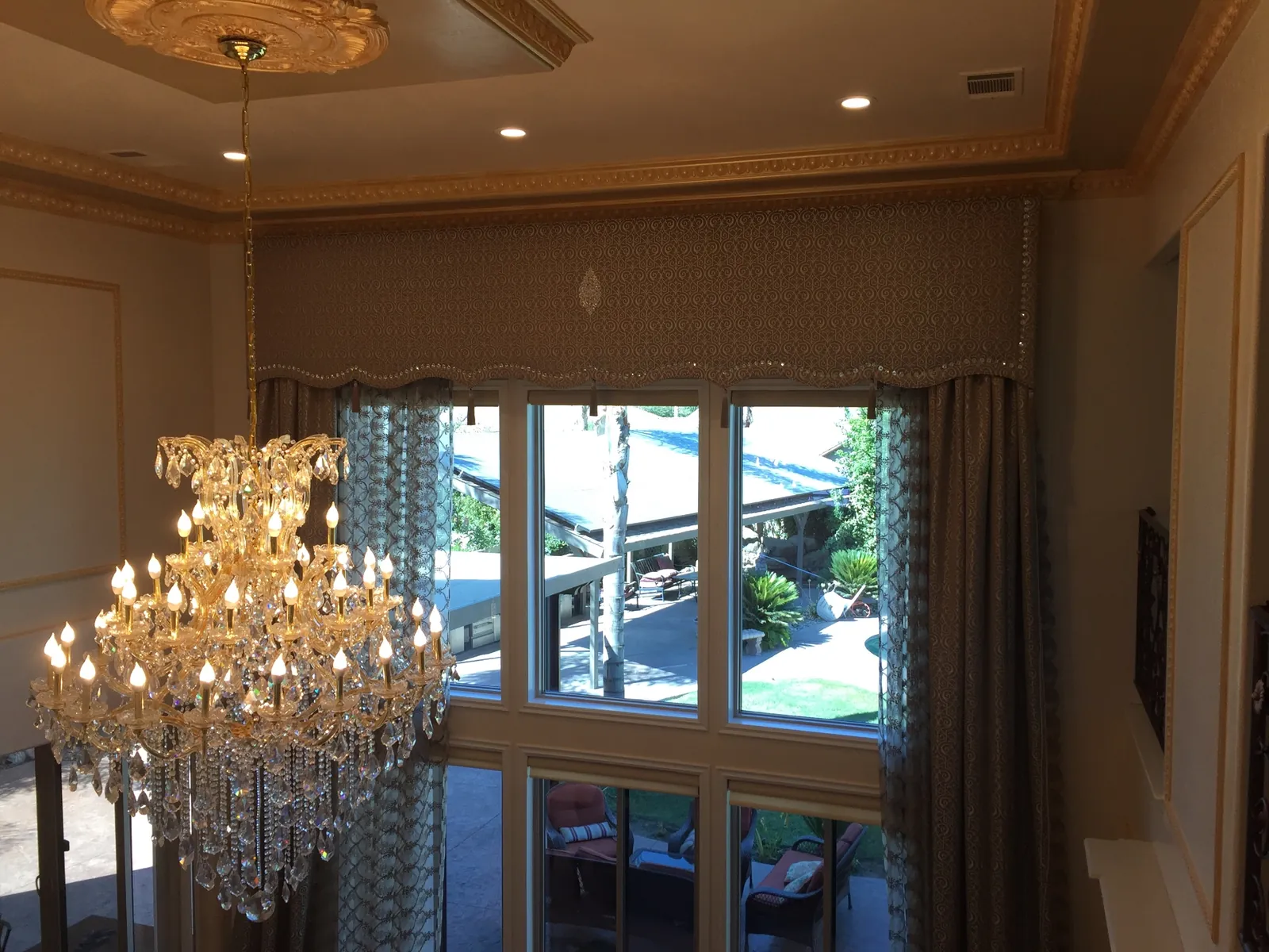 After — finished Porterville two-story living room drapery viewed from the upper landing, embellished damask cornice box with crystal bead trim and center medallion spans the tall window, patterned sheers and lined taupe drapery panels frame the backyard view under gilded crown molding and a Maria Theresa crystal chandelier