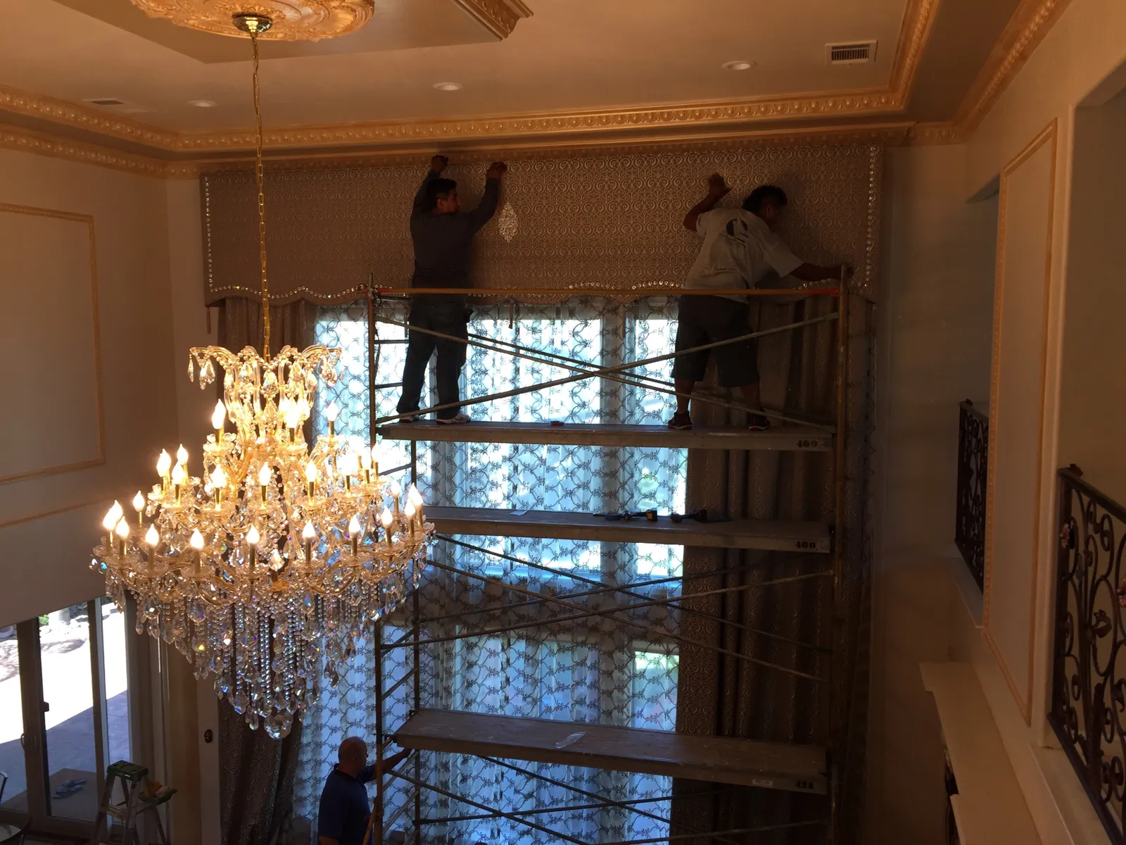 Wide view of the Porterville two-story living room during cornice install with two installers on scaffolding at ceiling height, large crystal chandelier in foreground, gilded crown molding above