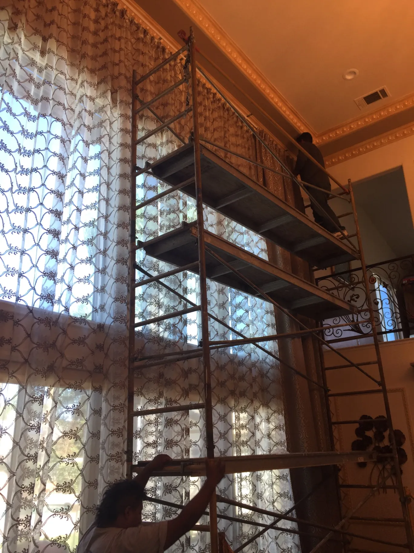 Two-story Porterville living room mid-install with scaffolding erected on the marble floor on floor-safe base plates, full-height patterned sheers already hung against the tall window wall