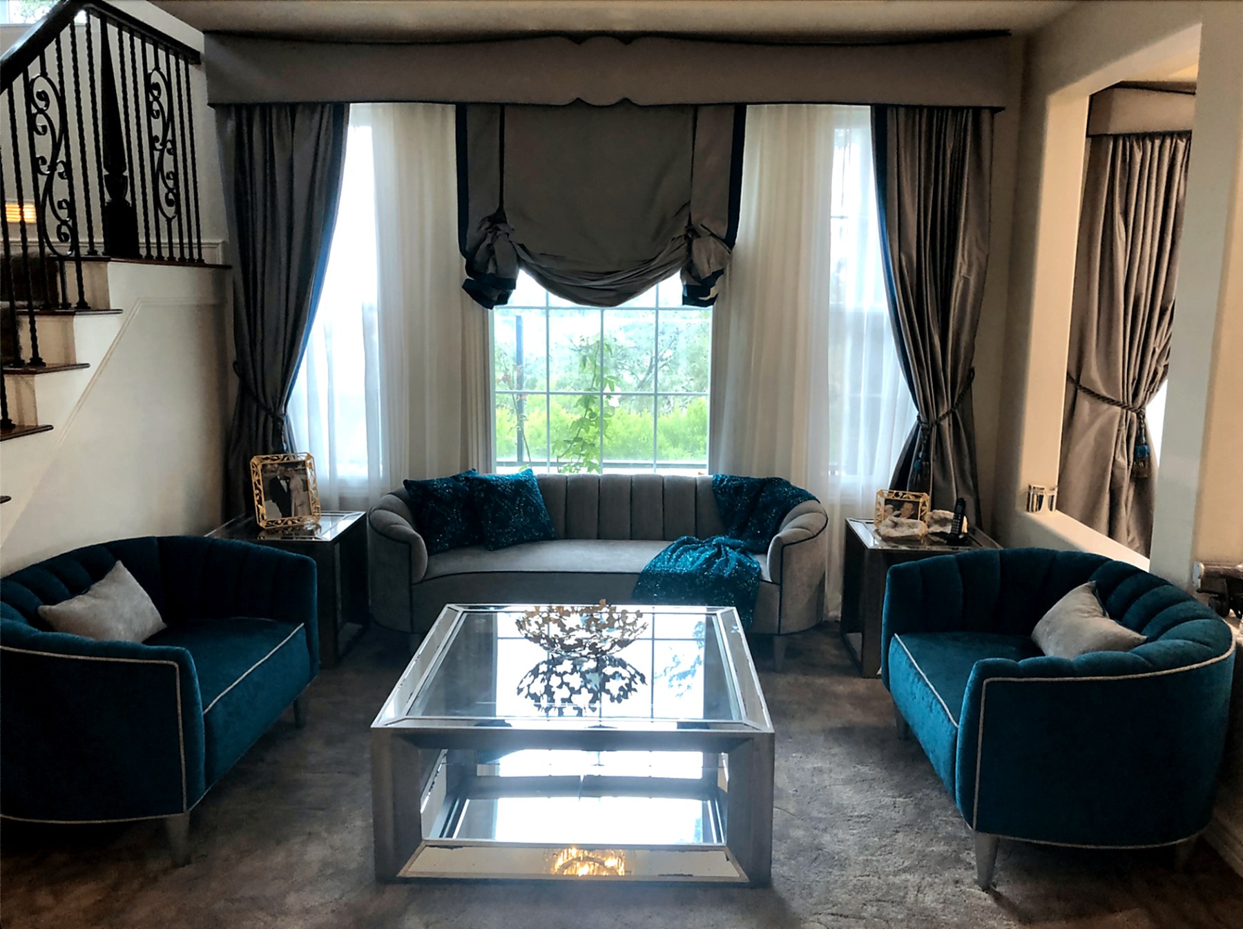 Laguna Niguel formal living room with a custom upholstered soft-gray cornice spanning the window wall, full-length silk drapery panels in gray with a turquoise leading edge over sheer under-panels, a relaxed Roman shade centered above a curved gray sofa flanked by teal velvet sofas and a mirrored coffee table