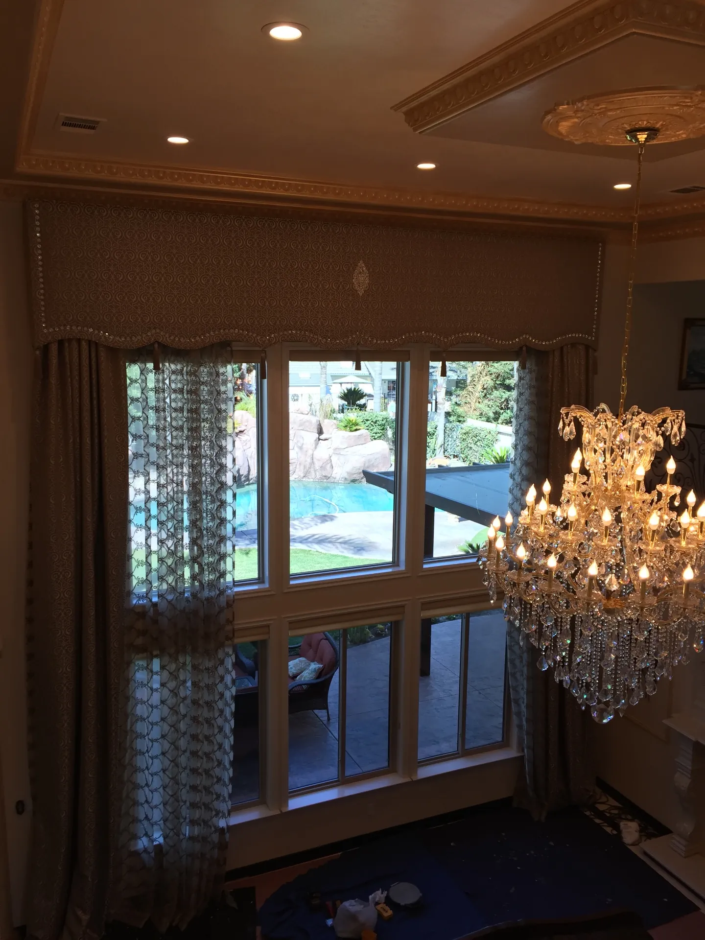 After — completed Porterville living room drapery from inside, shaped damask cornice with continuous crystal bead trim above patterned sheers and lined taupe side panels, pool, rock waterfall and lawn visible through the tall window, gilded ceiling molding and crystal chandelier