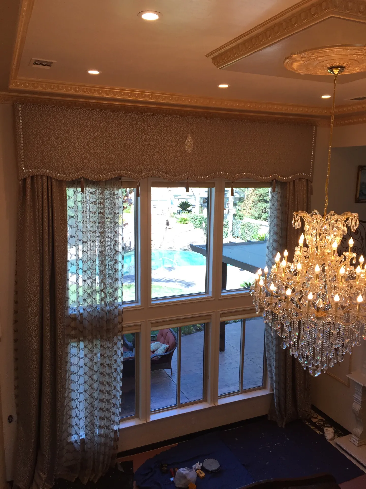 Completed Porterville living room window with embellished damask cornice box, crystal bead trim and center medallion, layered patterned sheer and warm-taupe lined drapery panels framing a tall pool-view window beneath gilded ceiling molding and a crystal chandelier