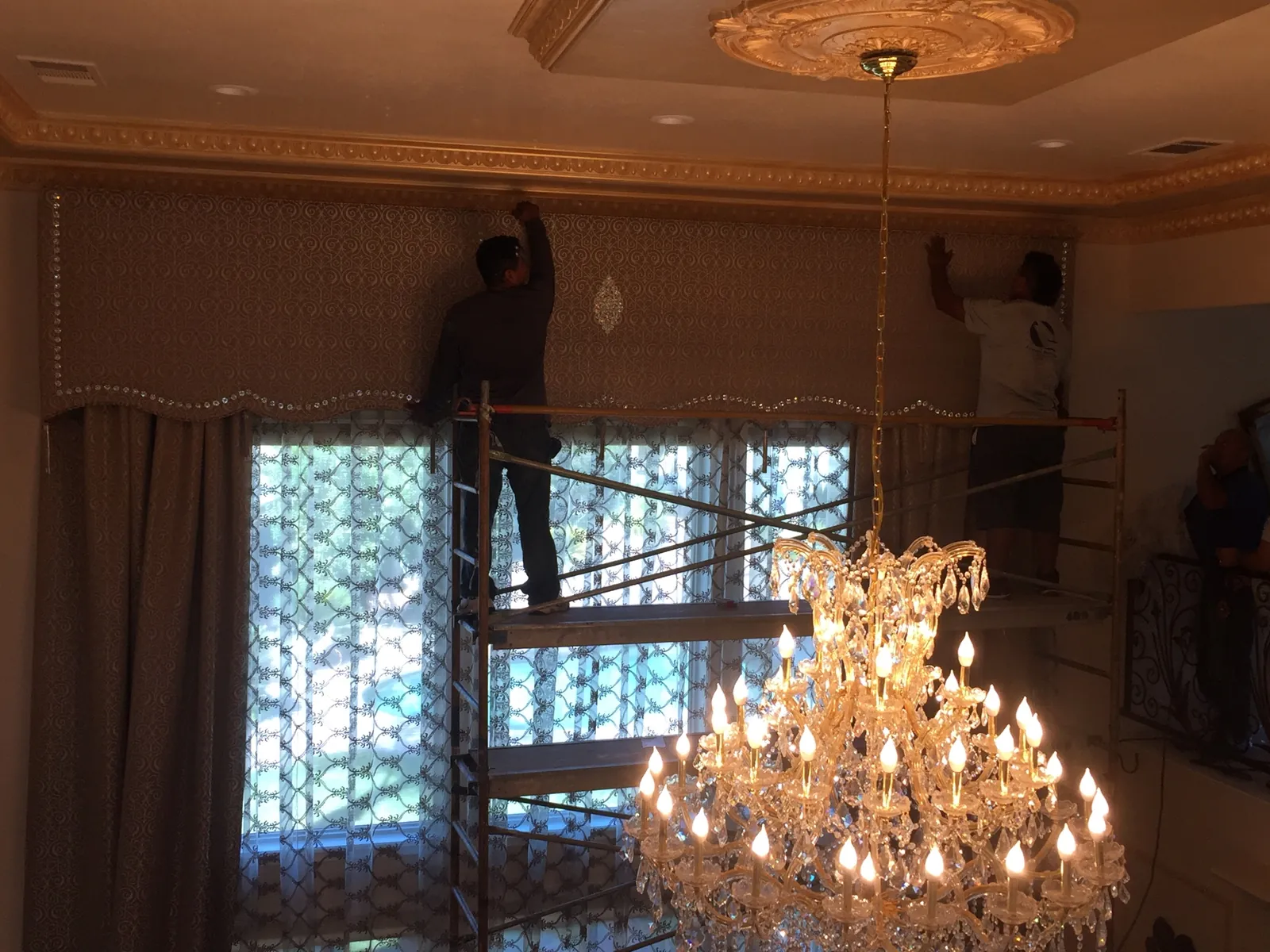 Two installers on scaffolding hand-setting the embellished damask cornice with crystal bead trim at the top of a tall Porterville living room window, gilded ceiling ornament above