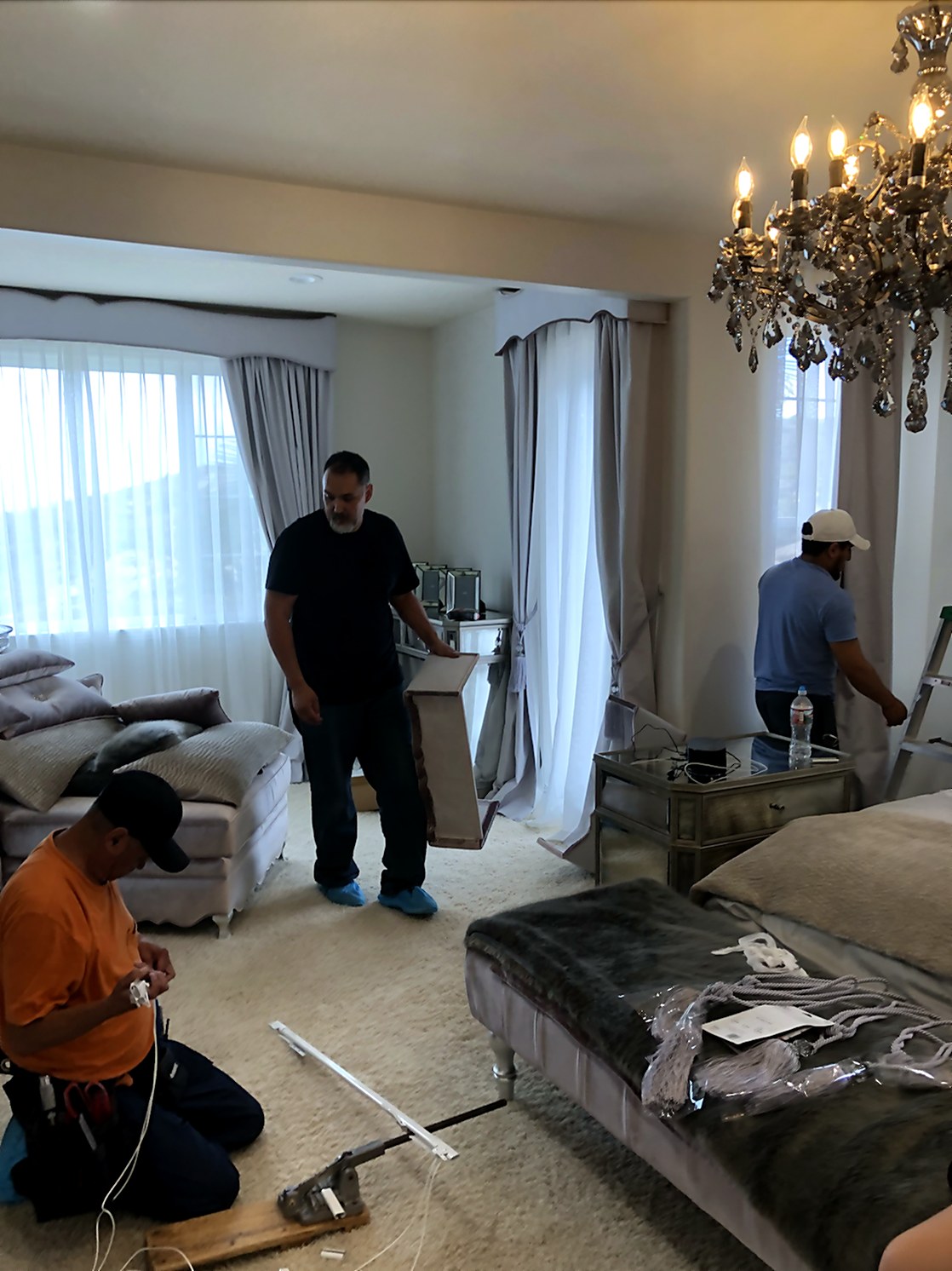 Master bedroom installation detail showing the soft-gray silk side panels with sheer under-layer being hand-dressed and steam-trained, with the workroom team setting hardware below a crystal chandelier