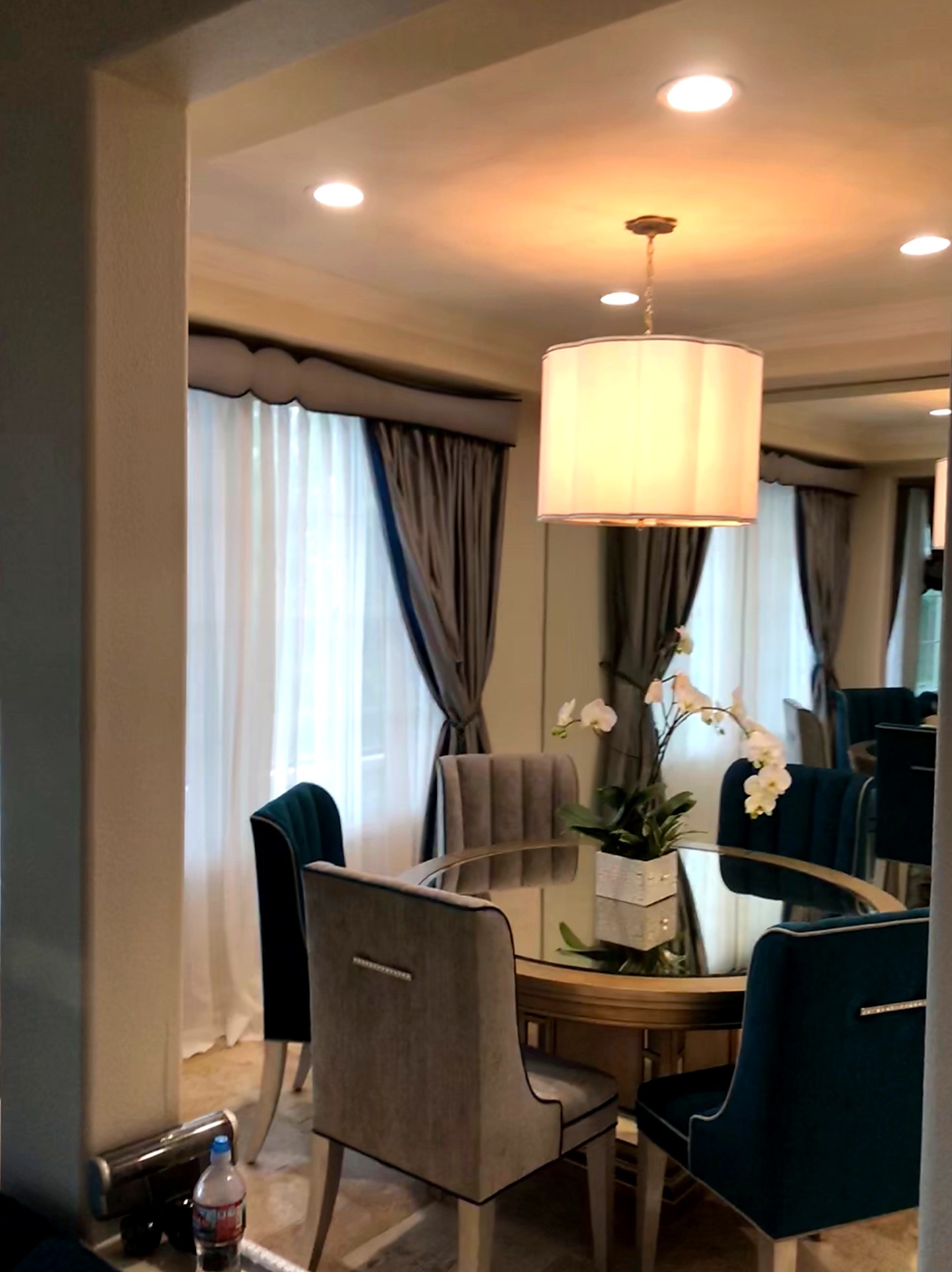 Laguna Niguel great room view through a cased opening showing scalloped upholstered cornice boxes in soft gray over silk drapery panels and sheer under-panels, set behind a round mirrored dining table with channel-tufted gray and teal velvet chairs