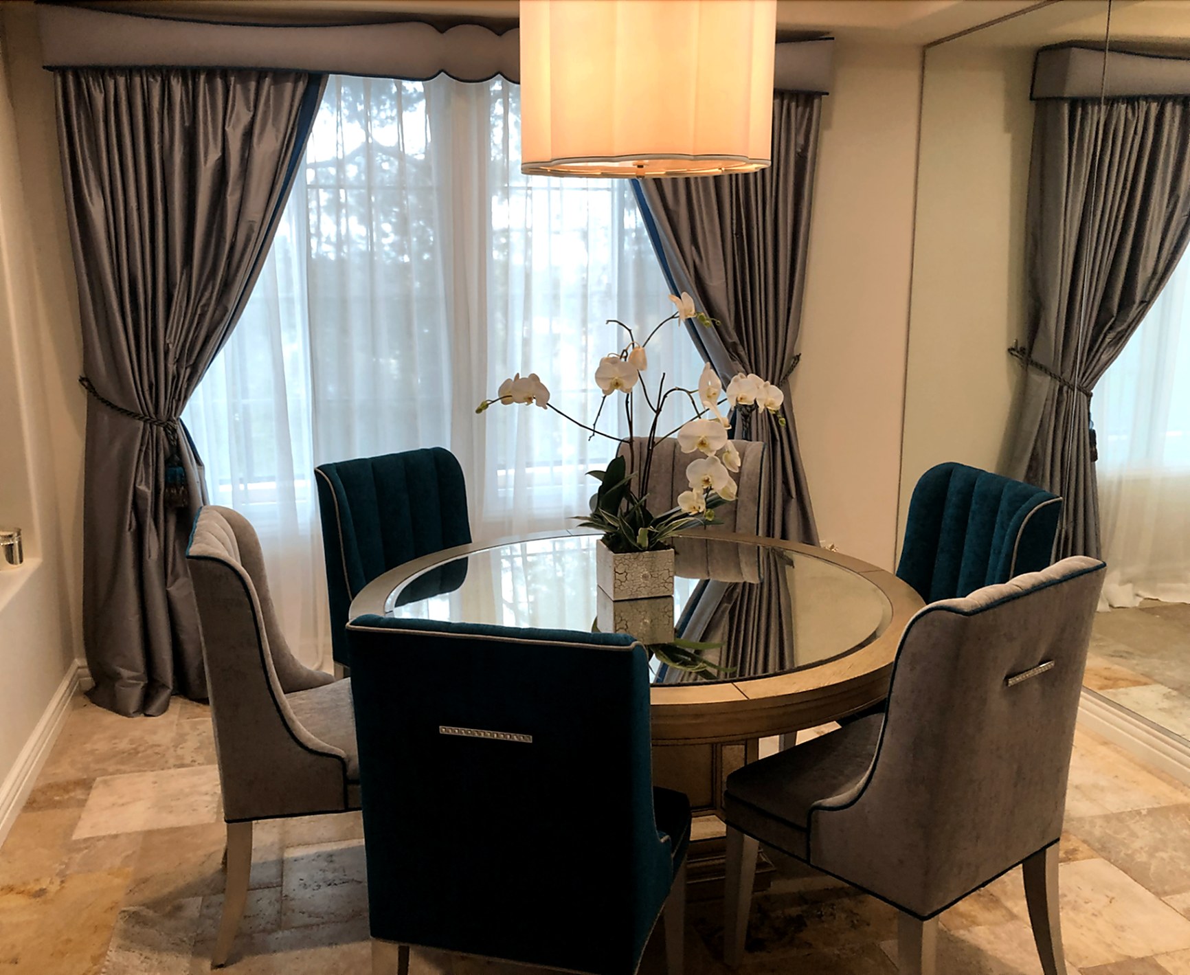 Master bedroom during installation: custom upholstered cornice in soft gray with scalloped lower edge, full-length silk drapery panels in soft gray over sheer under-panels framing tall bay windows, crystal chandelier overhead, installers steam-training panels and setting hardware