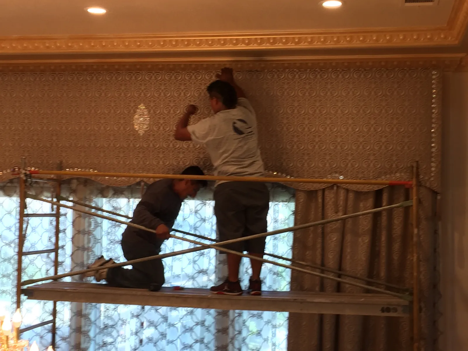 Close mid-install view of installers hand-tacking the embellished cornice box against the wall under heavy egg-and-dart gilded crown molding in the Porterville living room