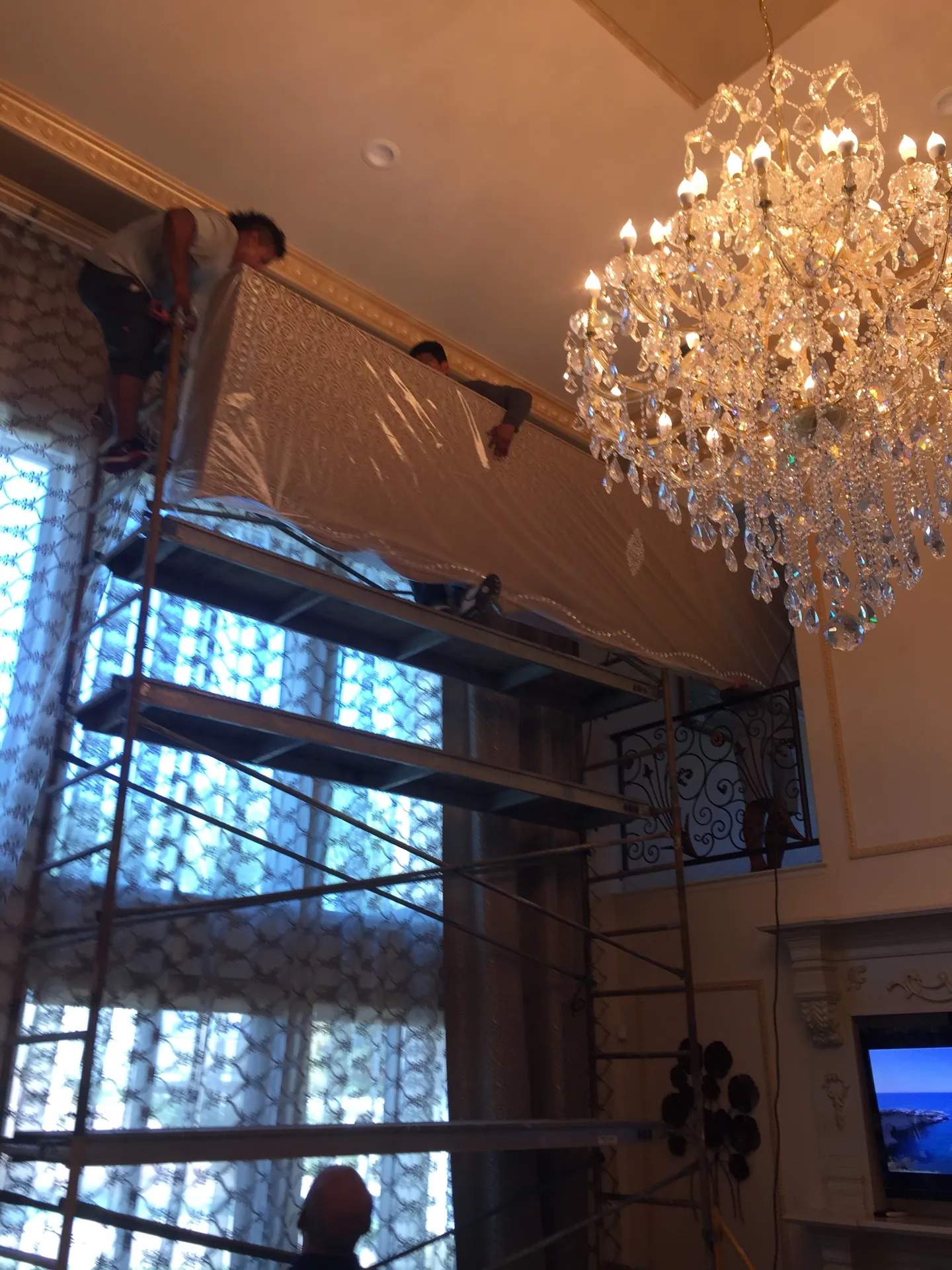 Installers lifting a long fabric-wrapped cornice box section in protective plastic up the scaffolding toward the gilded crown molding of the two-story Porterville living room