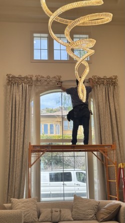 Side view of installer on scaffold positioning ornate cast-brass top treatments along the header of a tall arched window beneath a large crystal chandelier