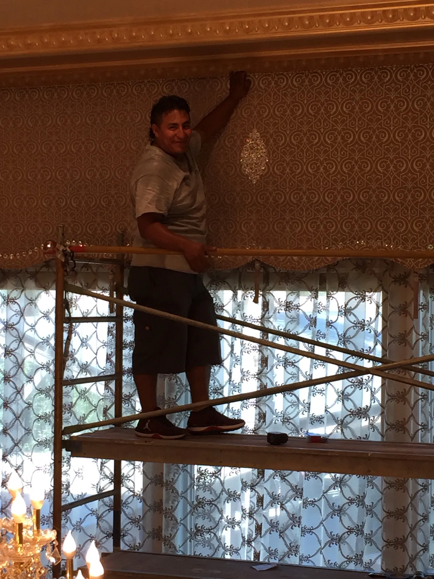 Smiling installer on scaffolding finishing the upper edge of the patterned damask cornice box against the gilded molding of the Porterville living room