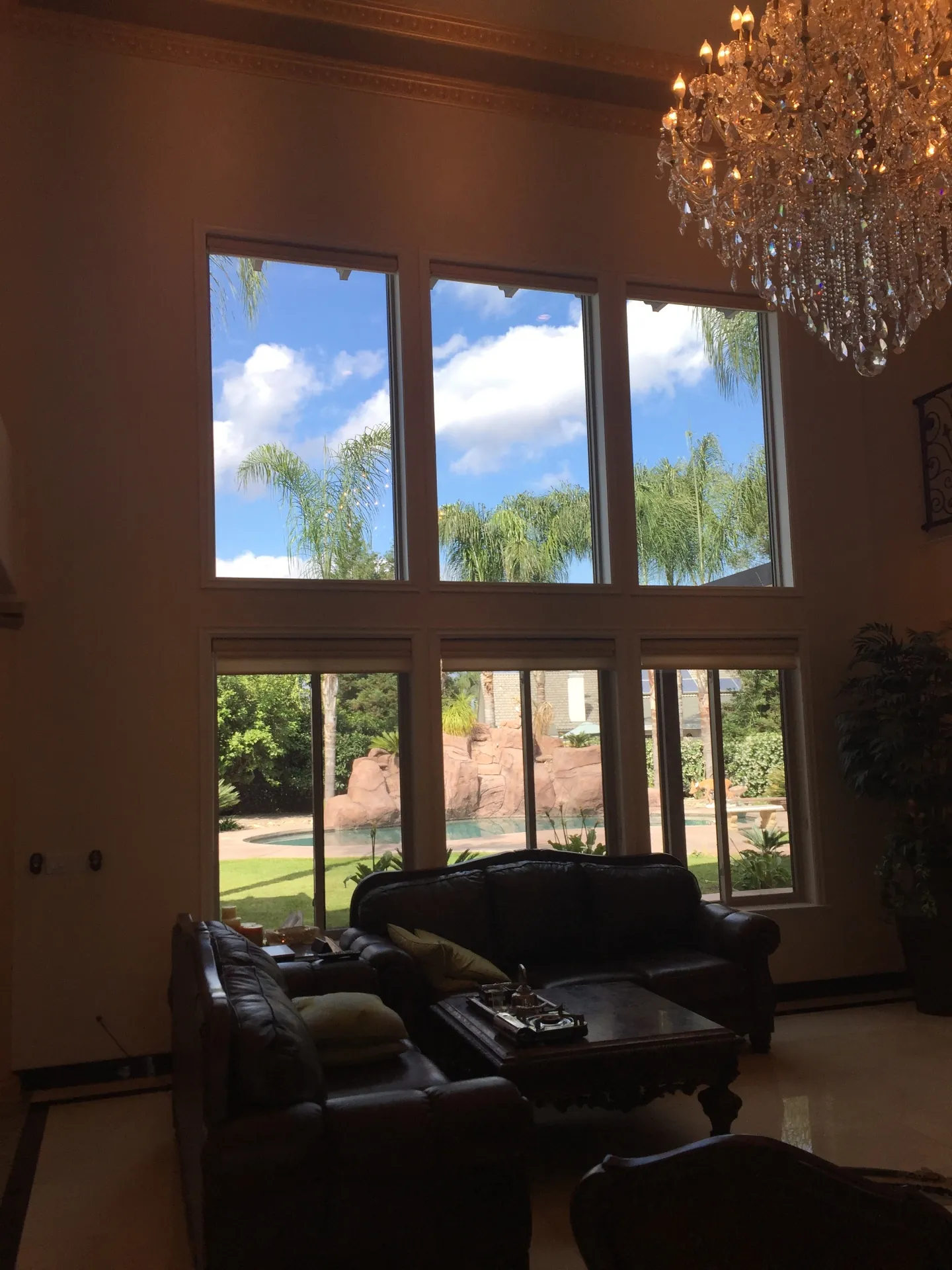Before — Porterville two-story living room window wall completely bare, six tall windows showing pool, palm trees and bright sky, dark leather sofas, ornate crystal chandelier and gilded crown molding above, no drapery or cornice yet installed